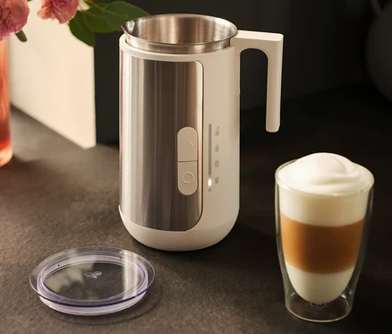 Spieniacz do mleka ze stali nierdzewnej stoi obok szklanki kawy latte i pokrywki na ciemnym blacie.