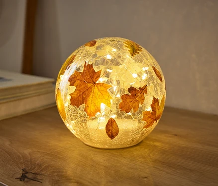 Kulista lampa o popękanej szklanej fakturze, ozdobiona jesiennymi liśćmi, ciepło świeci na drewnianej powierzchni. Małe światełka w środku oświetlają liście.