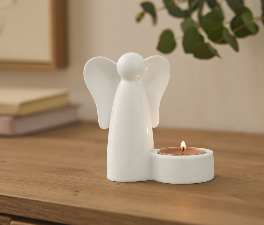 Biały ceramiczny świecznik w kształcie anioła z zapaloną świeczką typu tealight na drewnianej powierzchni. W tle rozmyte książki i roślina.