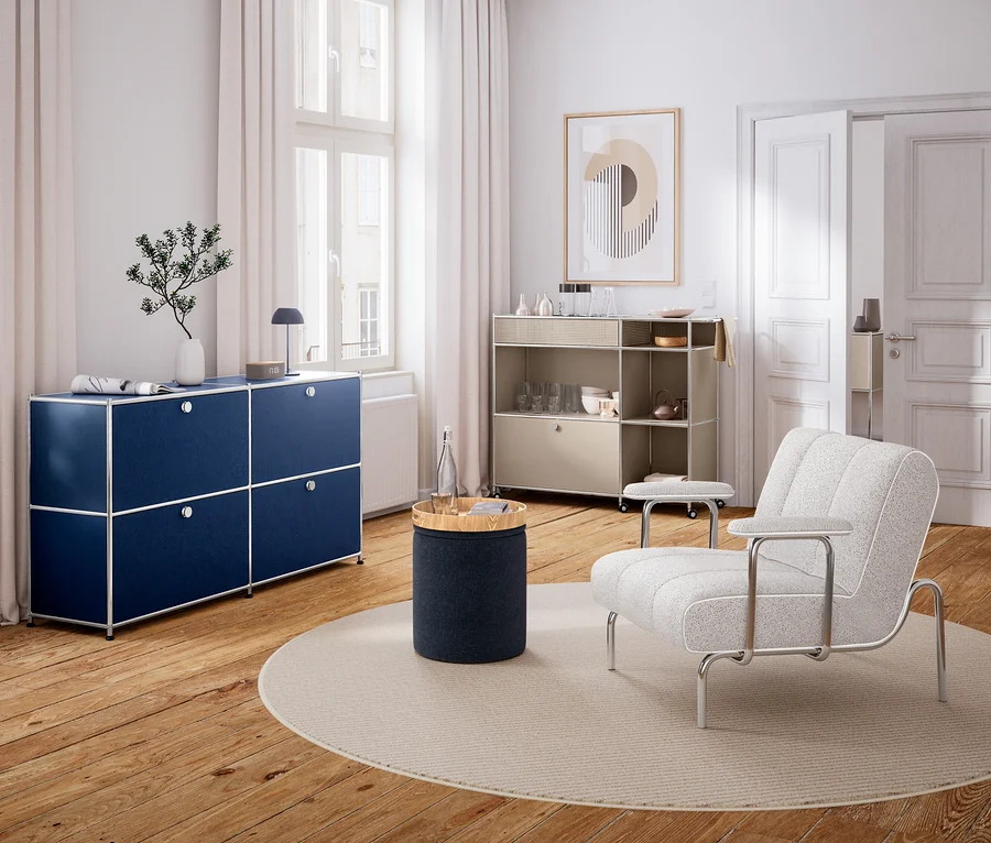 Nowoczesny salon z drewnianą podłogą i białymi ścianami. Po lewej stronie, pod oknem, stoi granatowa komoda modułowa z wazonem i lampą. Na środku okrągły dywan z cylindrycznym stolikiem bocznym i butelką wody. Po prawej stronie jasny fotel i beżowy regał na kółkach z naczyniami. Na ścianie wisi abstrakcyjny obraz.