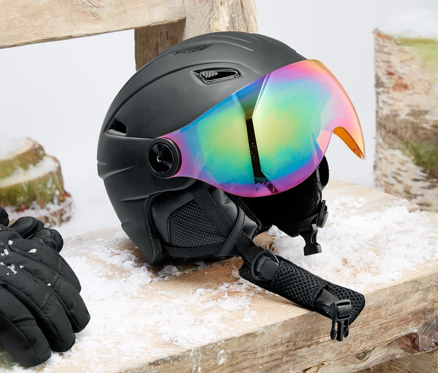 Kask narciarski i snowboardowy typu hardshell z wizjerem leży na drewnianej ławce. Obok leżą czarne rękawice.