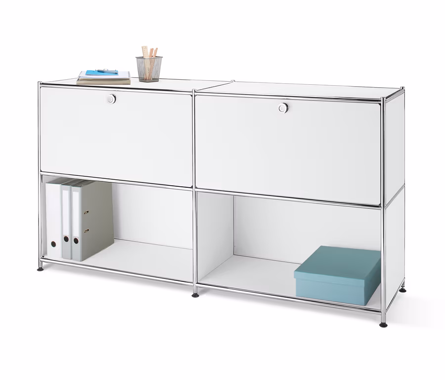 Widok z przodu metalowej szafki typu sideboard z 2 przegrodami z klapami, montowanymi w dowolnym miejscu, na której leży notatnik i szklanka z długopisami. W otwartej półce znajdują się dwa segregatory i niebieskie pudełko.