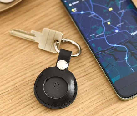Na stole leżą klucze z czarnym inteligentnym lokalizatorem z breloczkiem oraz telefon komórkowy z mapą.