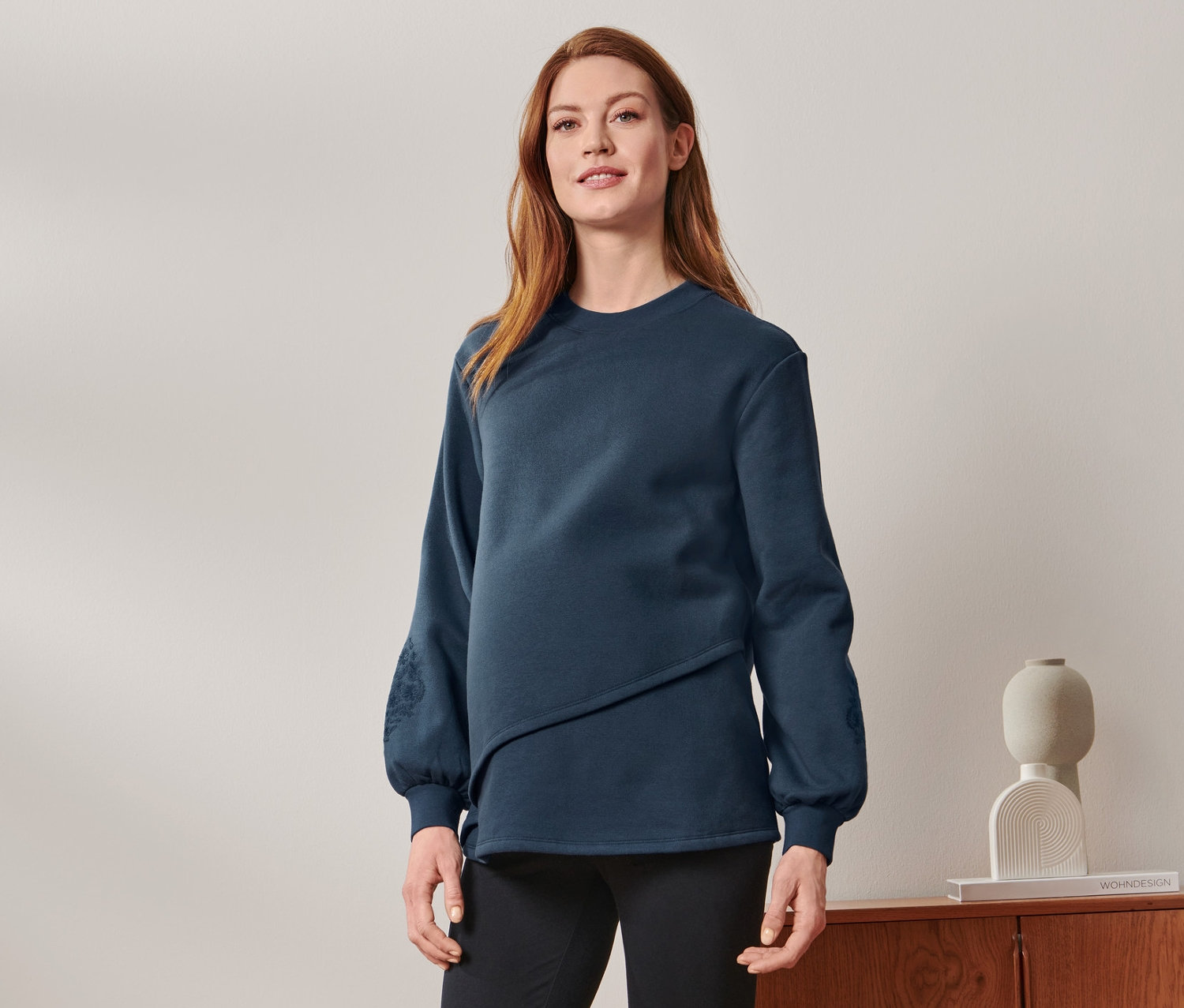 Ciężarna kobieta stoi, ubrana w bluzę ciążową i do karmienia, czarne spodnie i tenisówki.