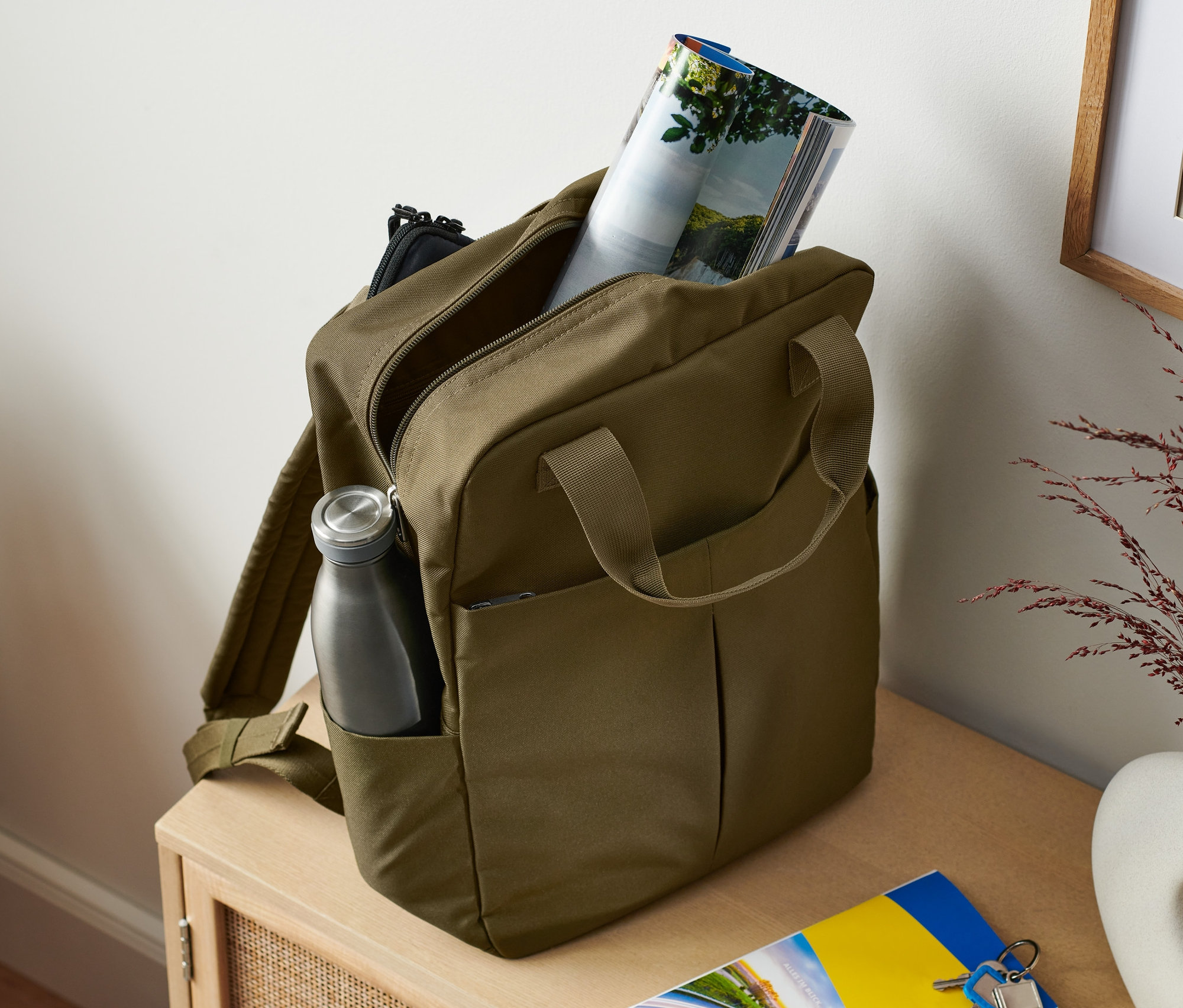 Plecak z zabezpieczonymi paskami w kolorze khaki z butelką wody w bocznej kieszeni i czasopismem w środku.