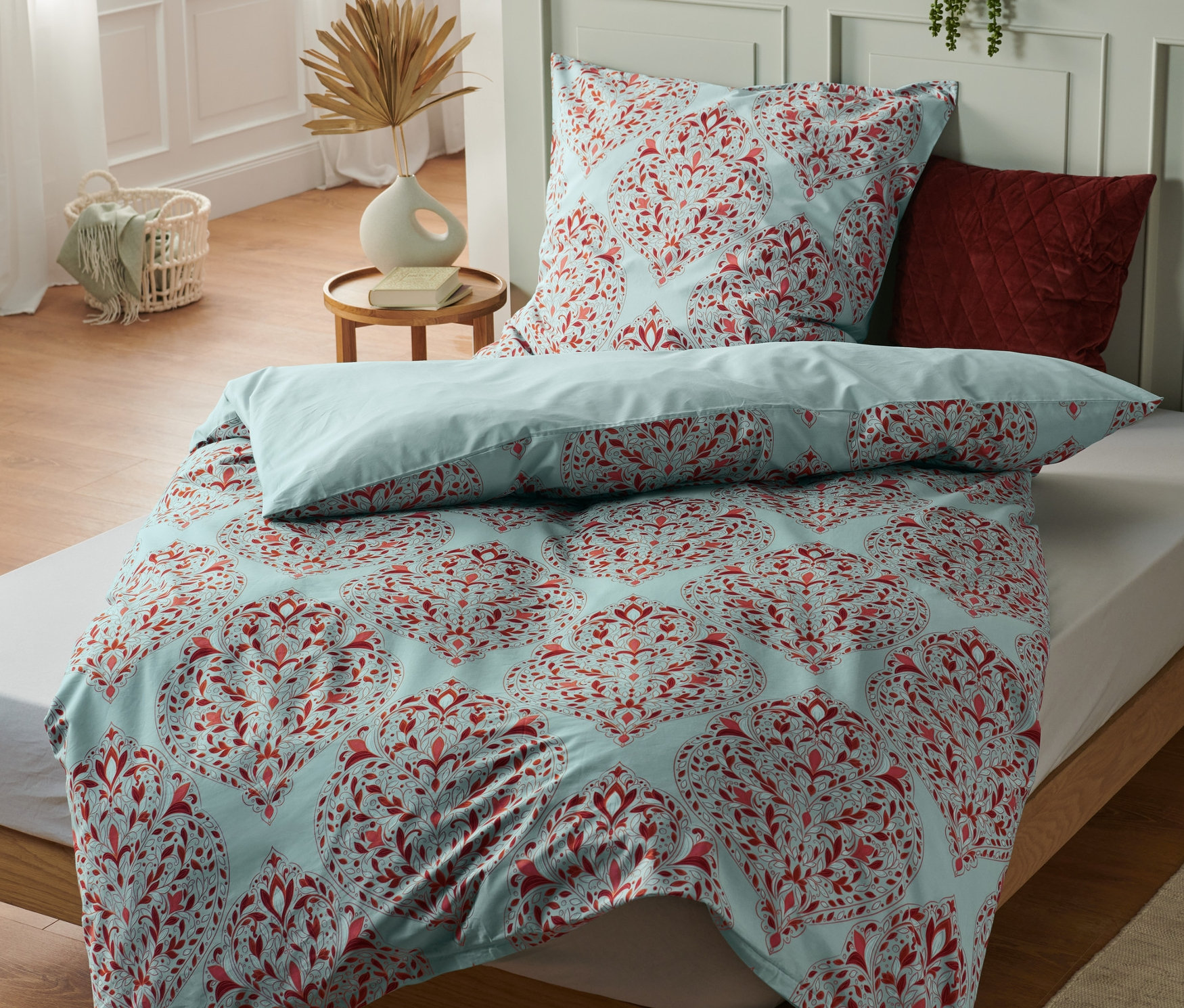 Łóżko z niebieską pościelą w czerwone kwiaty. Na łóżku leżą trzy komplety pościeli bawełnianej premium, wielkość standardowa.