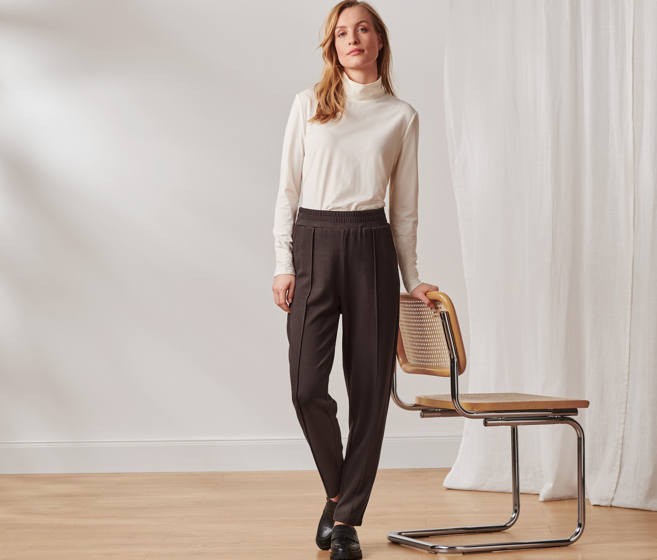Kobieta stoi obok krzesła, ma na sobie ciemnobrązowe spodnie dresowe z zakładką.