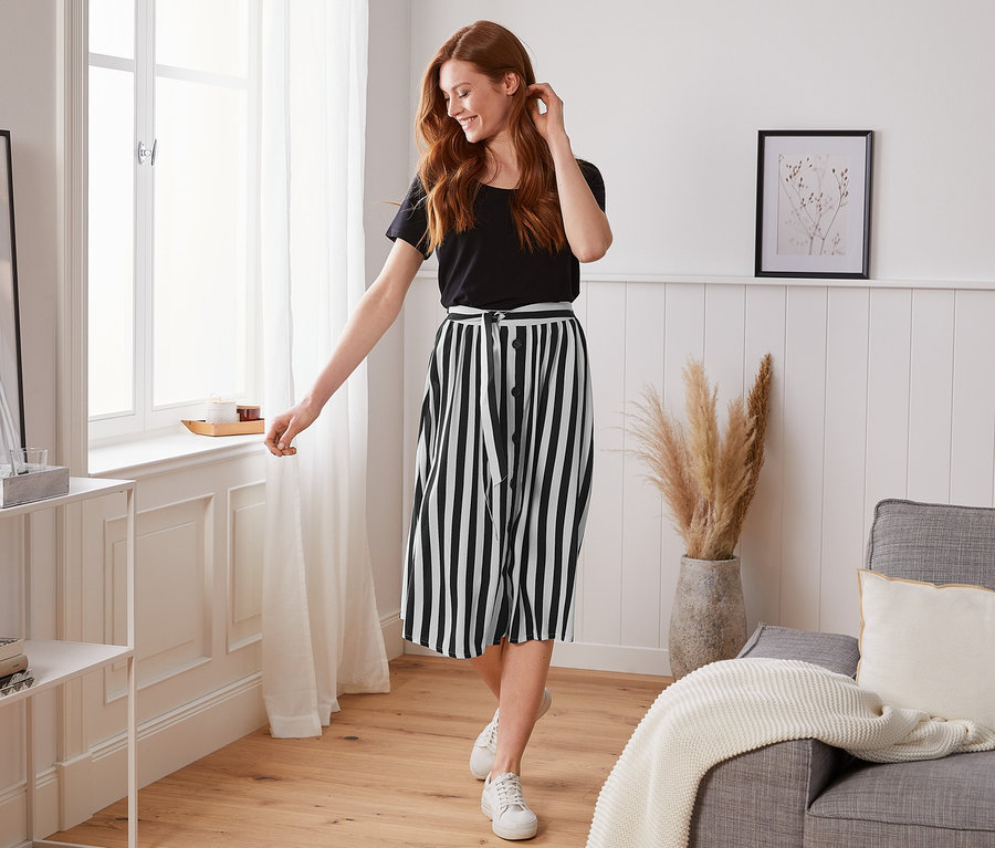 Uśmiechnięta rudowłosa kobieta pozuje w czarnym T-shircie i pasiastej spódnicy z tkaniny.