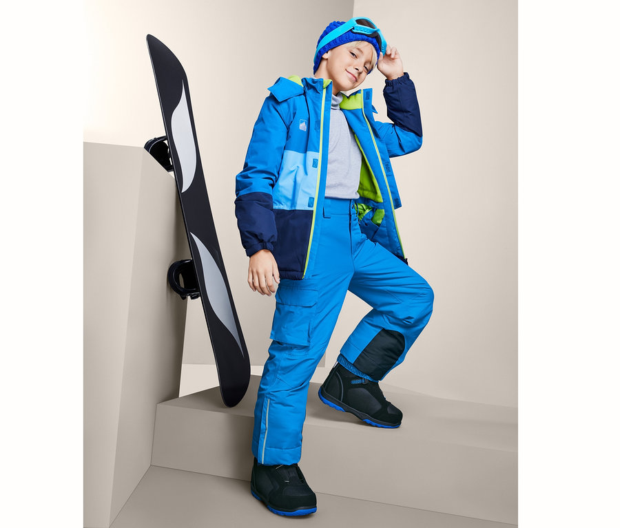 Chłopiec w niebieskich spodniach snowboardowych stoi obok deski snowboardowej.