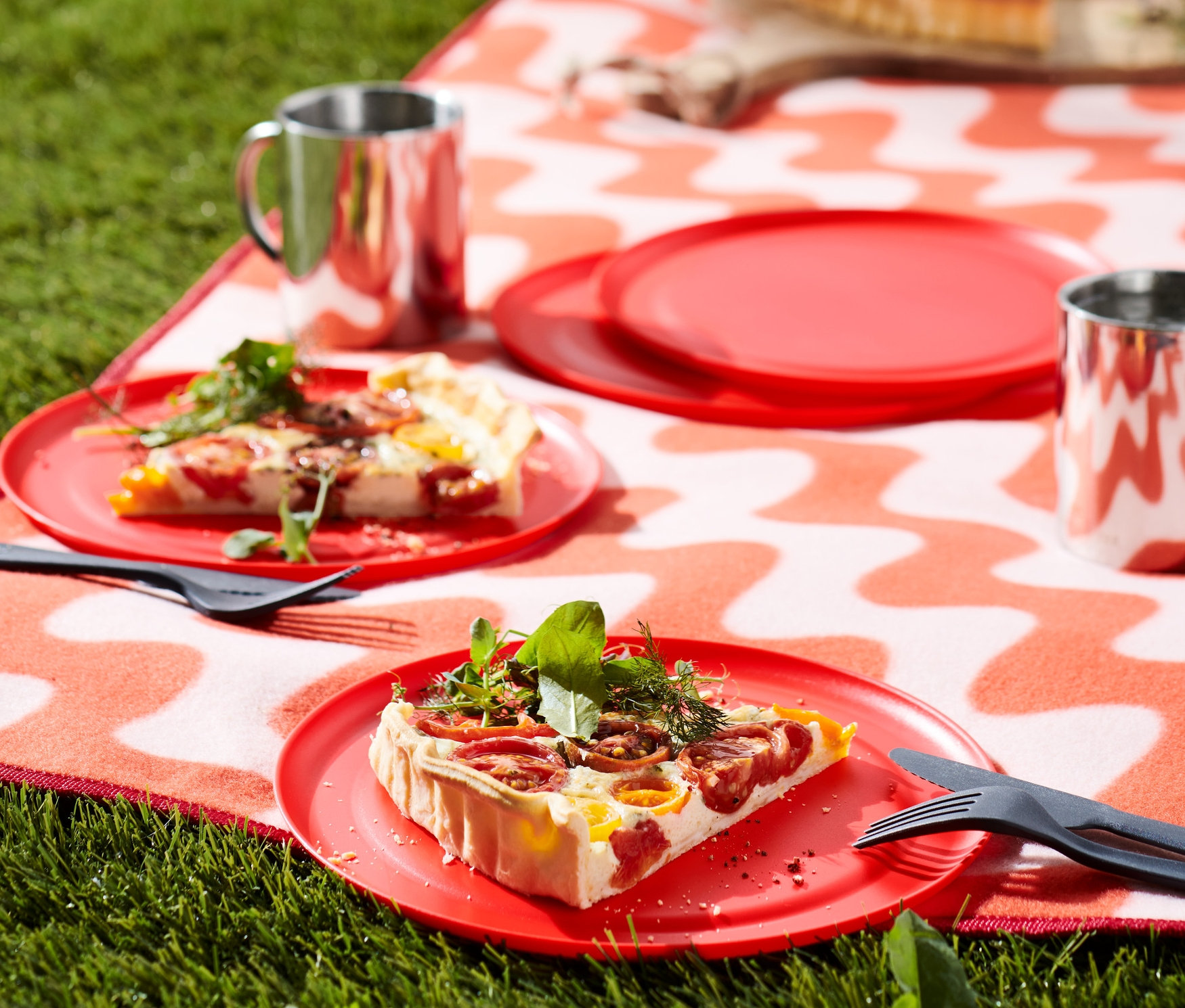 Na trawie leży koc we wzór falisty. Na kocu znajdują się talerze piknikowe, 4 sztuki z jedzeniem, dwa kubki i sztućce. W tle plecak i dodatkowe jedzenie.