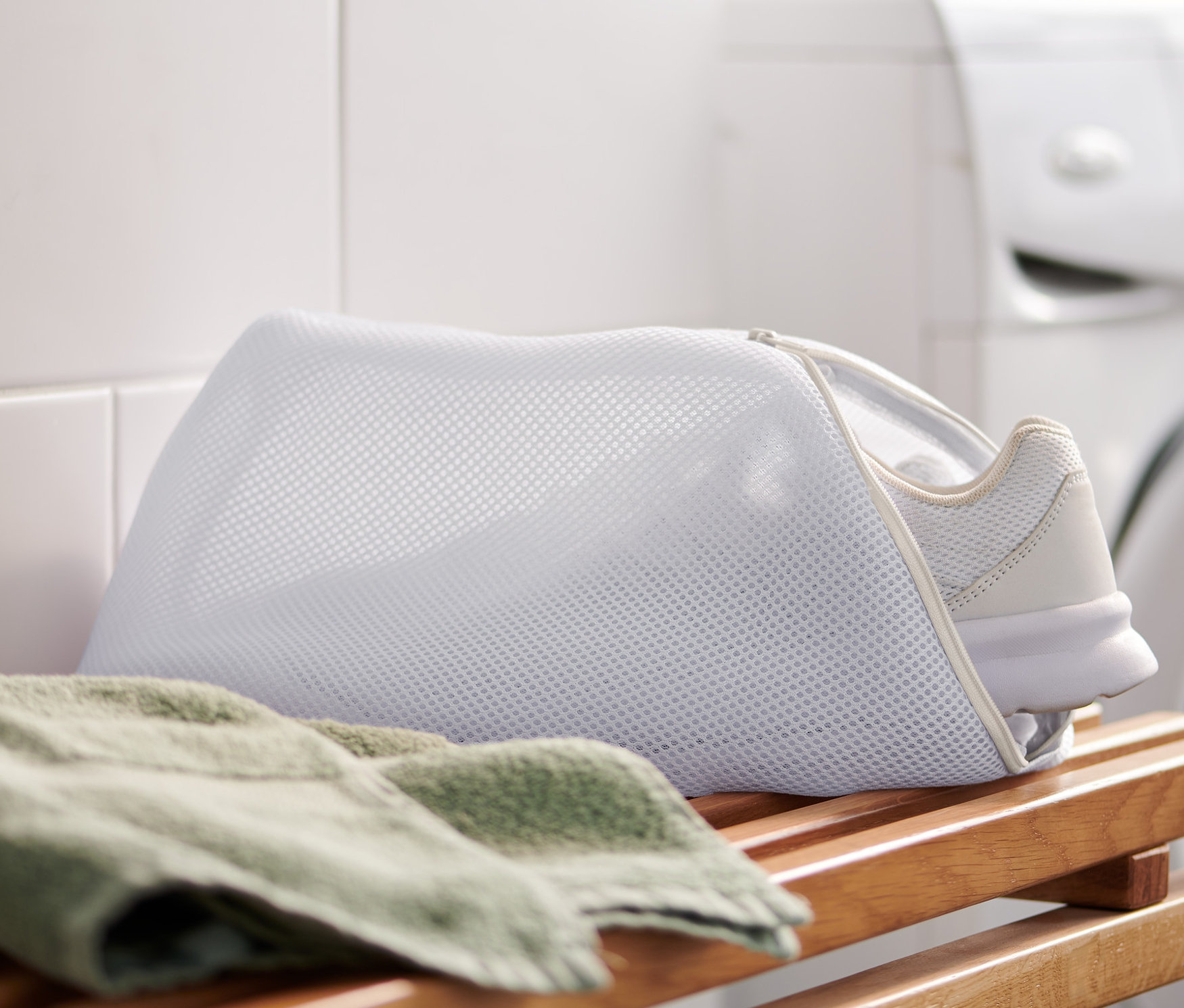 Biały but sportowy umieszczony jest w woreczku do prania butów, który leży na drewnianej ławce obok zielonego ręcznika. W tle widoczna jest biała pralka i białe płytki.