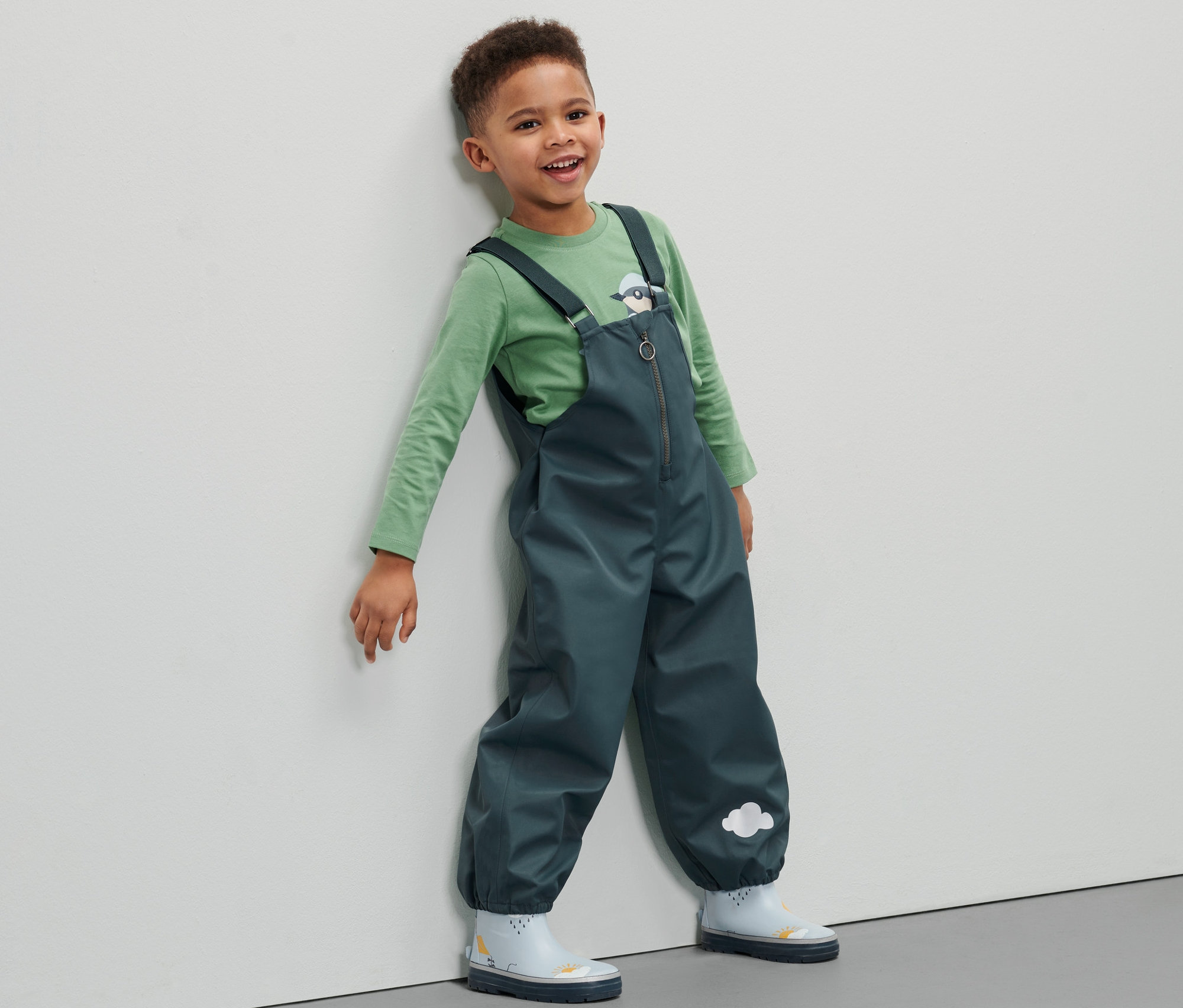 Chłopiec w spodniach przeciwdeszczowych opiera się o ścianę.