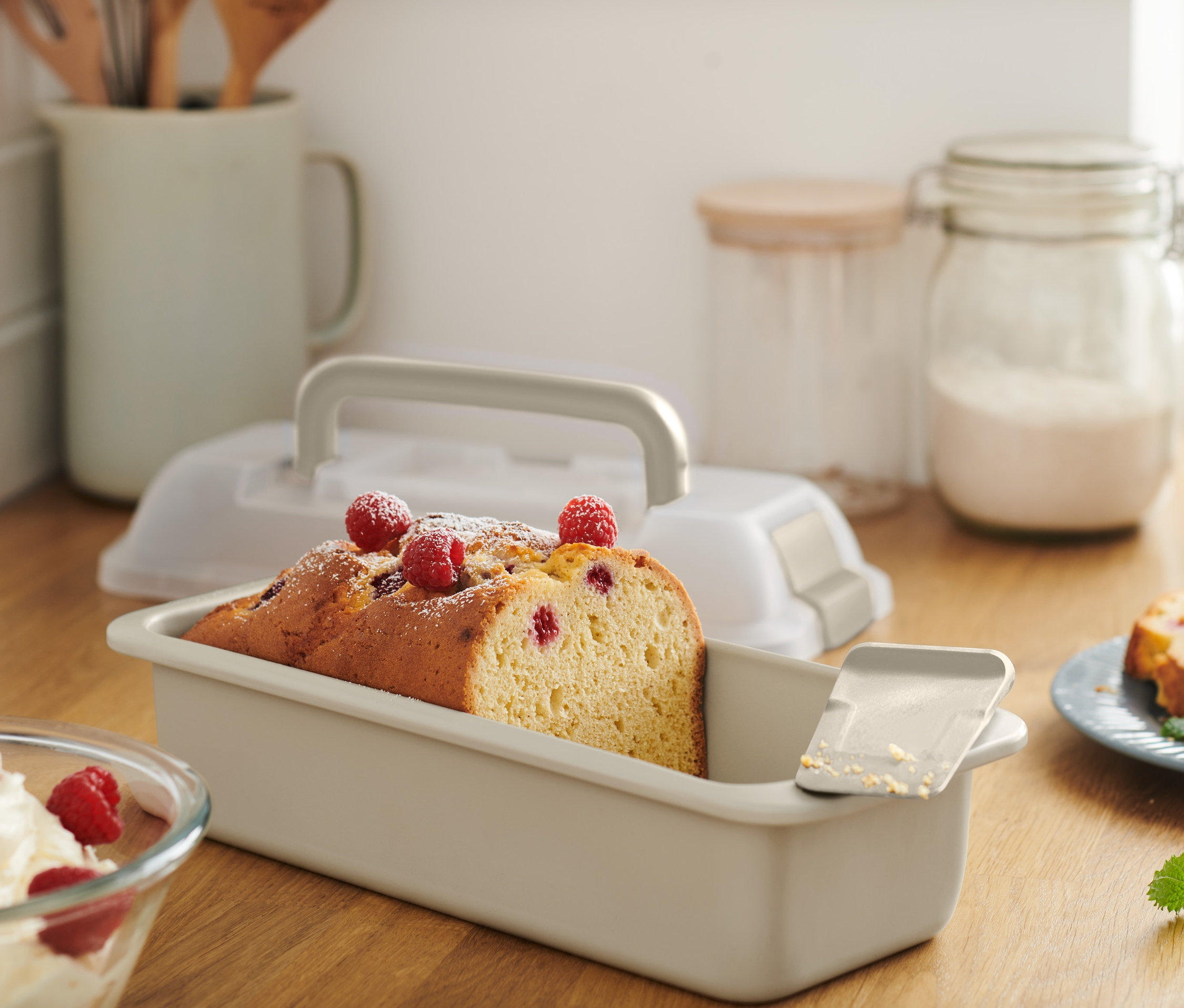 Pokrojone ciasto w prostokątnej formie do pieczenia z pokrywką do transportu, posypane cukrem pudrem i malinami.