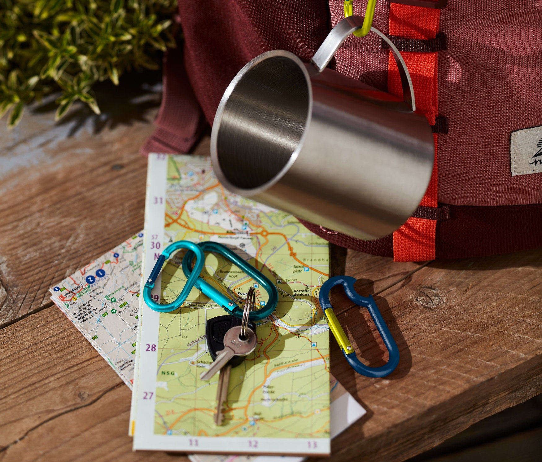Kubek ze stali nierdzewnej wisi na plecaku za pomocą jednego z 4 karabińczyków. Na mapie leżą klucze z kolejnym karabińczykiem.