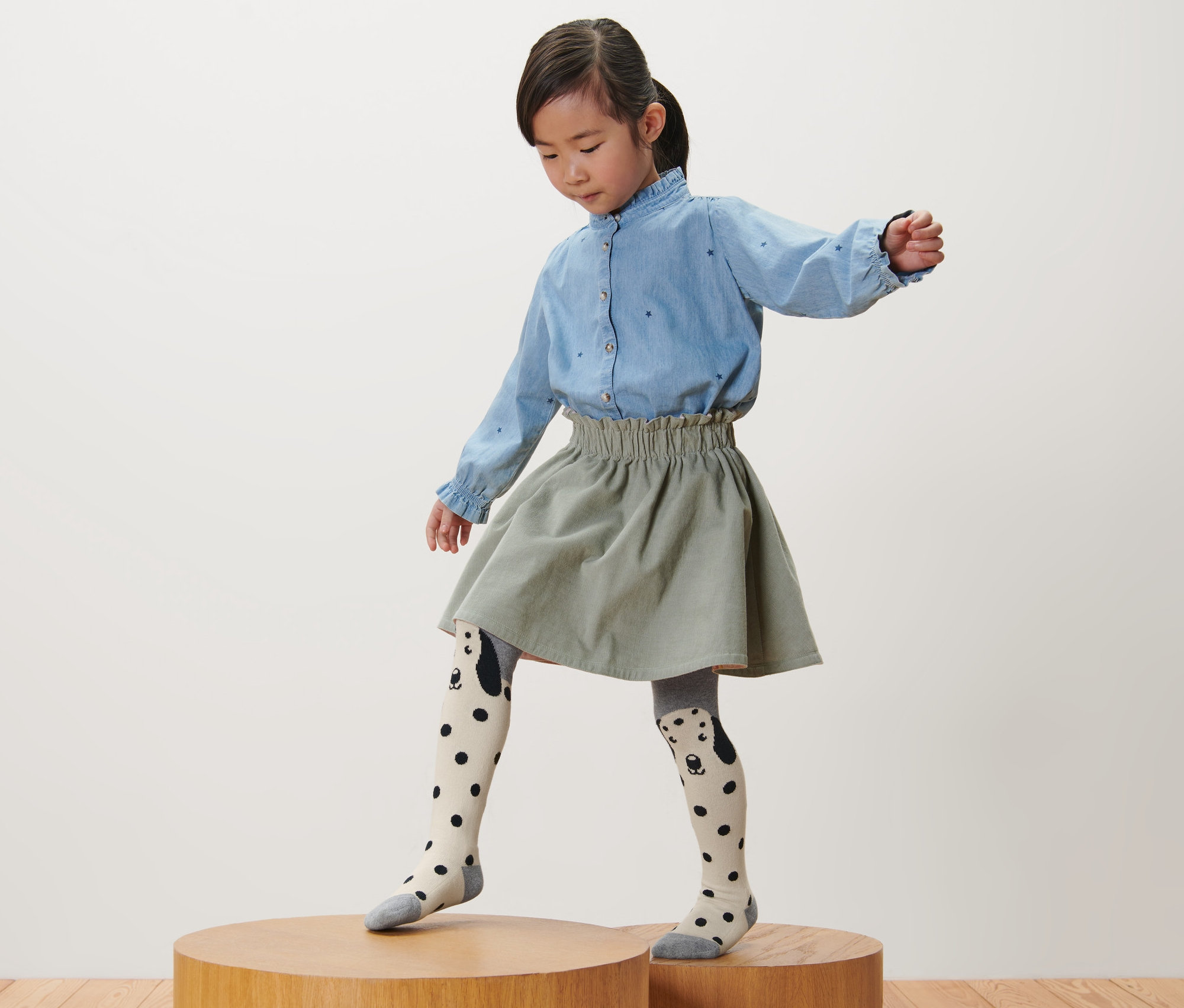 Dziewczynka stoi na drewnianym postumencie, ubrana w różowe, szare i jagodowe rajstopy dziecięce, 3 pary.