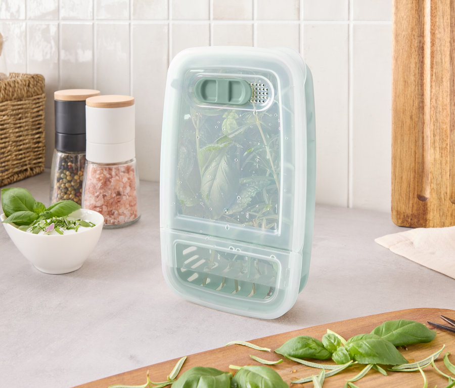 Pojemnik do przechowywania ziół z ziołami w środku stoi na blacie obok składników i deski do krojenia.