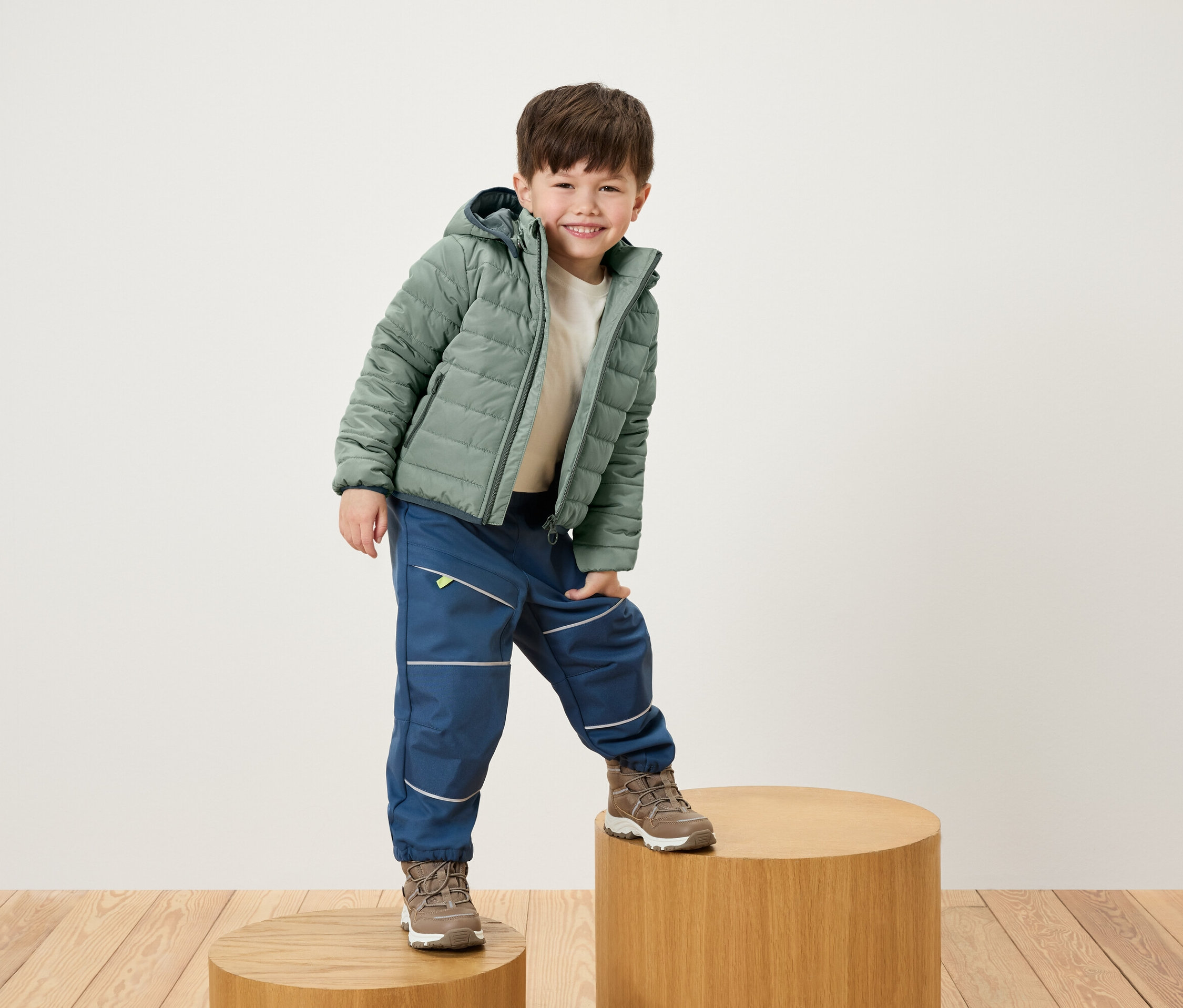 Uśmiechnięty chłopiec pozuje na drewnianym cokole w zielonej kurtce, niebieskich spodniach i brązowych butach.