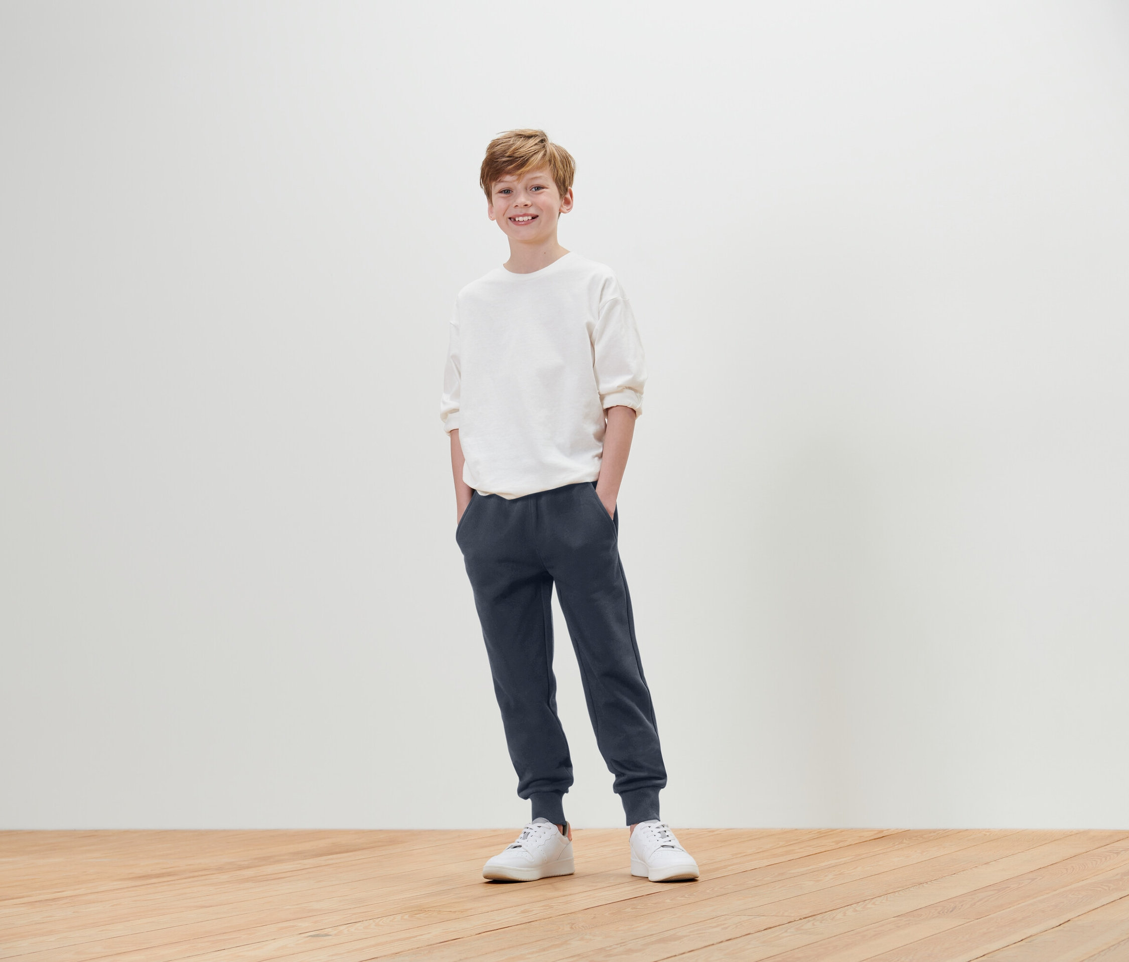 Chłopiec stoi z rękami w kieszeniach, ubrany w białą koszulkę, niebieskie spodnie dresowe i białe tenisówki.