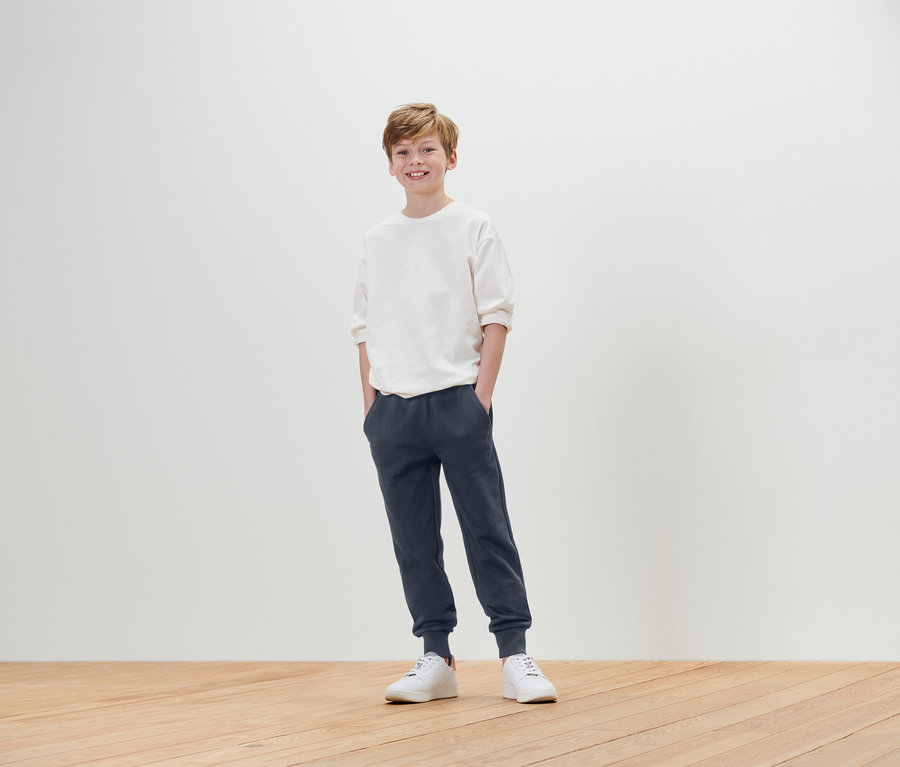 Chłopiec stoi z rękami w kieszeniach, ubrany w białą koszulkę, niebieskie spodnie dresowe i białe tenisówki.