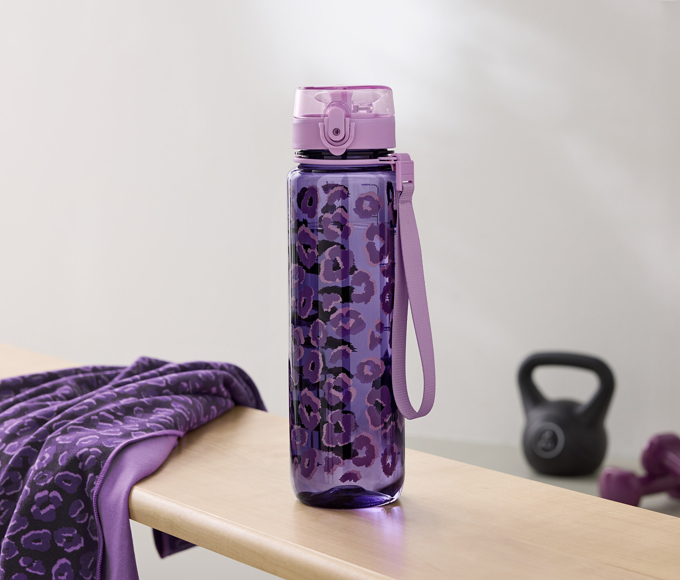 Fioletowa butelka na wodę we wzory leży obok ręcznika i hantli na ławce.