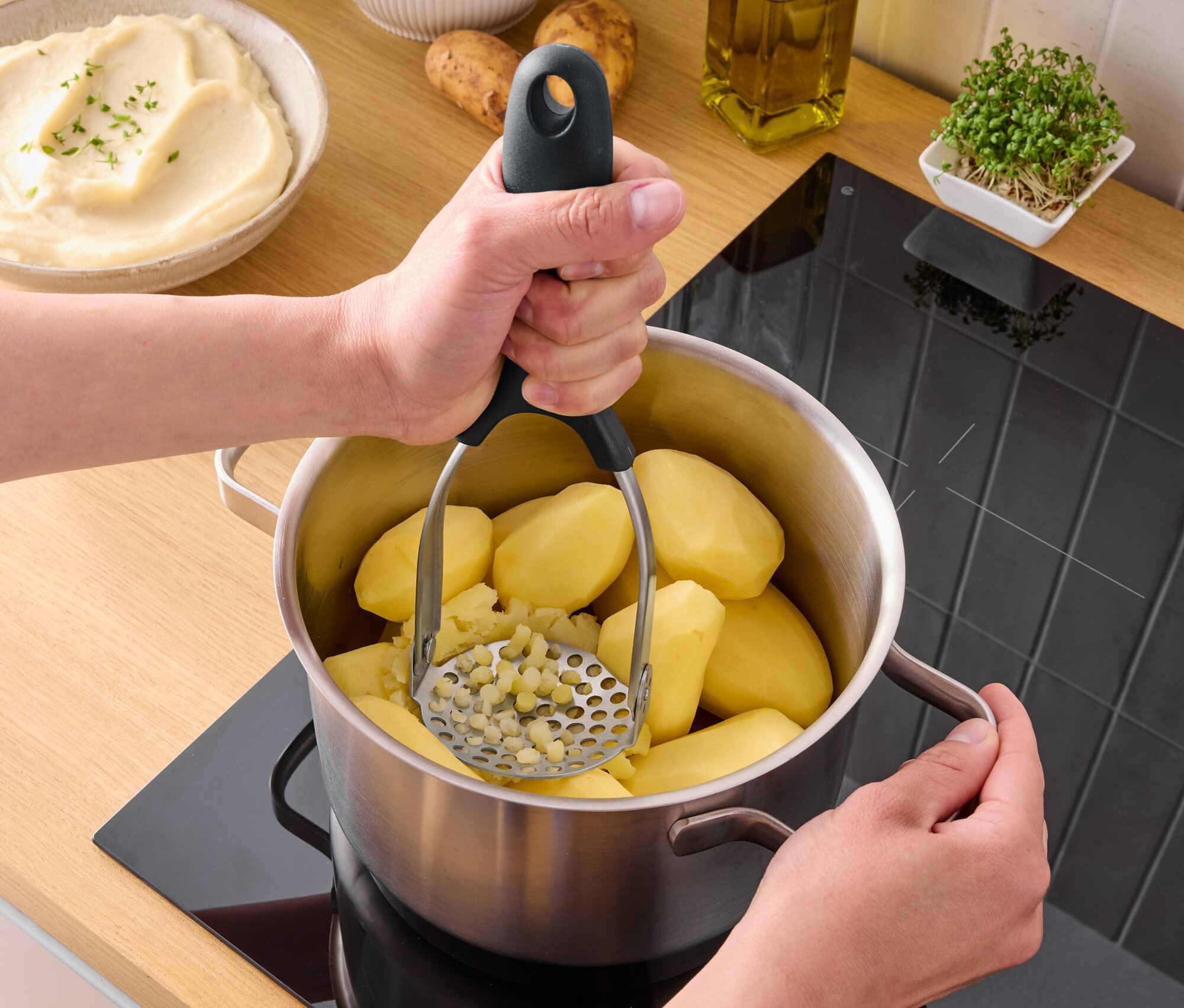 Osoba tłucze gotowane ziemniaki w garnku na kuchence za pomocą tłuczka do ziemniaków.