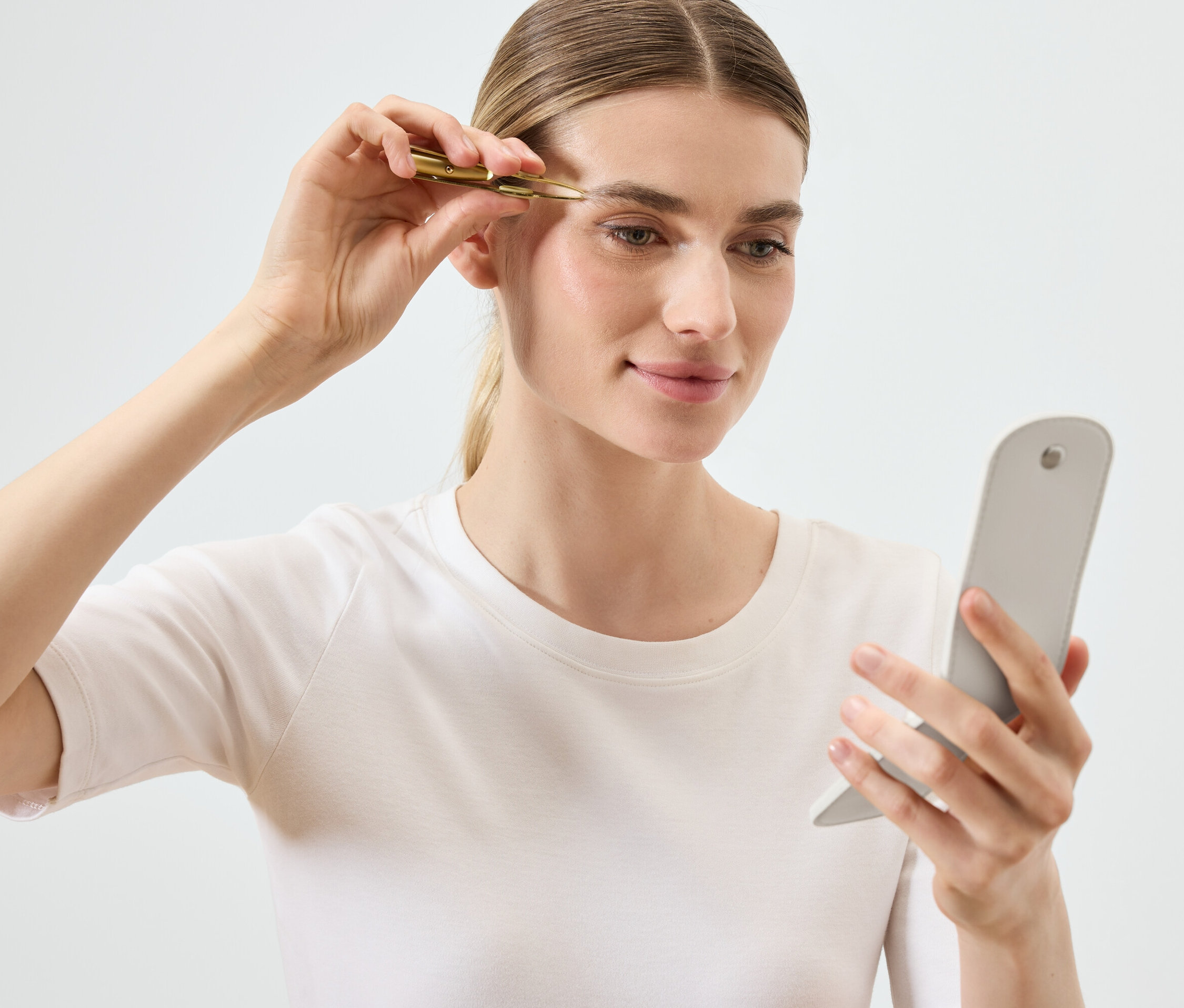 Kobieta reguluje brwi pęsetą, patrząc w lusterko.