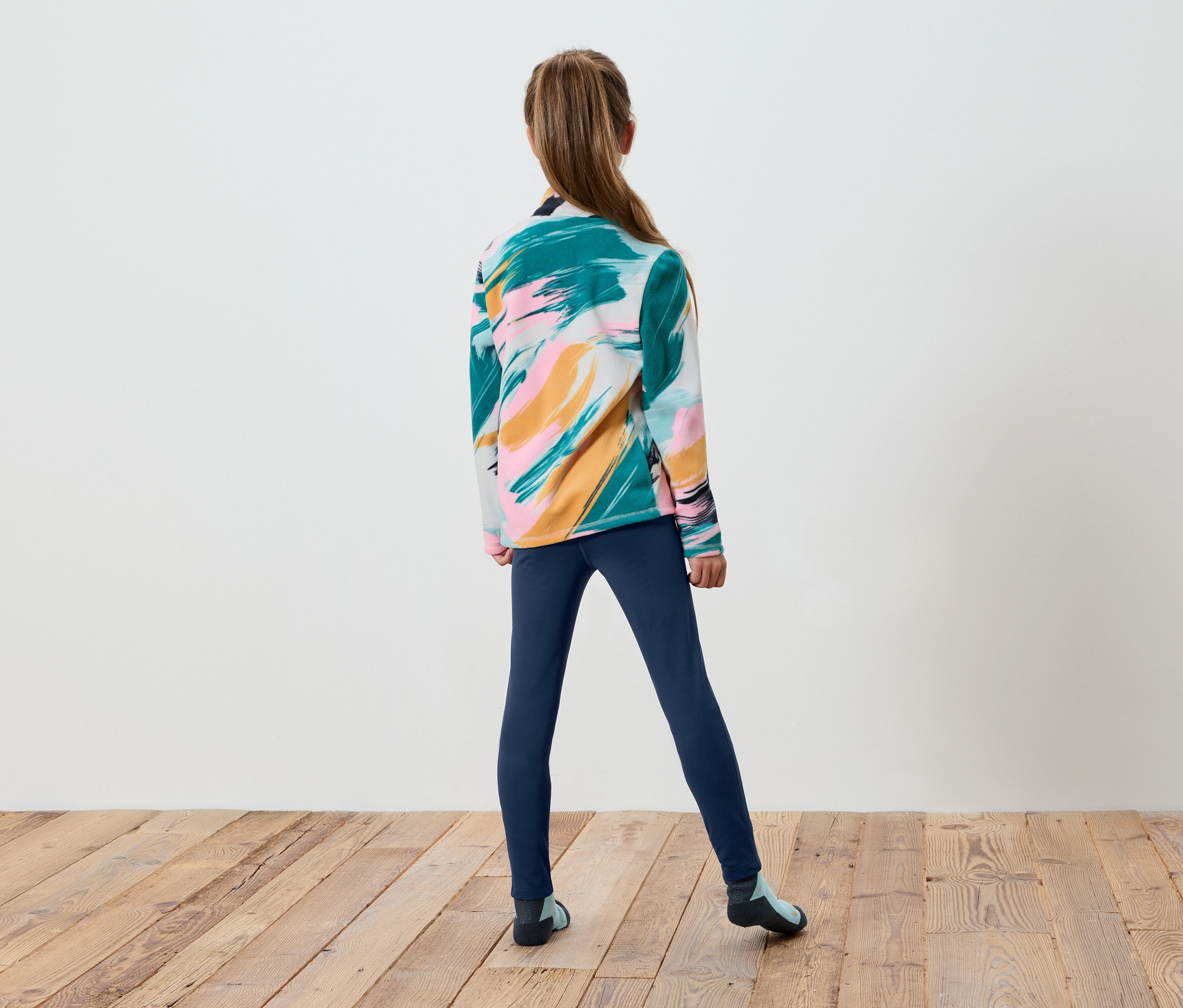 Dziewczyna stoi tyłem, ma na sobie kolorowy sweter i niebieskie legginsy.