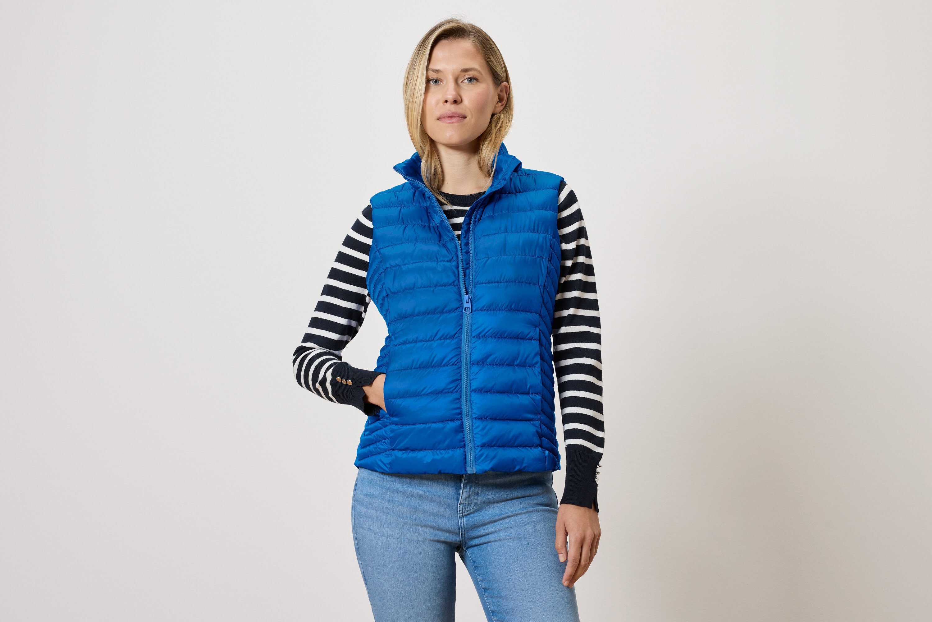 Kobieta w niebieskiej pikowanej kamizelce na zamek błyskawiczny, swetrze w czarno-białe paski i niebieskich dżinsach.