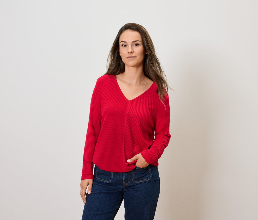 Kobieta z brązowymi włosami pozuje w czerwonym swetrze z długim rękawem i niebieskich dżinsach.