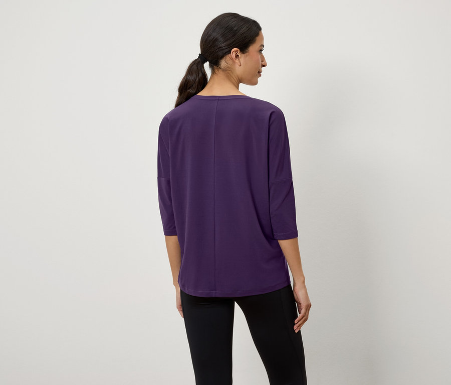 Kobieta z ciemnymi włosami związanymi w kucyk stoi tyłem, ubrana w fioletową koszulkę z rękawami trzy czwarte i czarne legginsy.