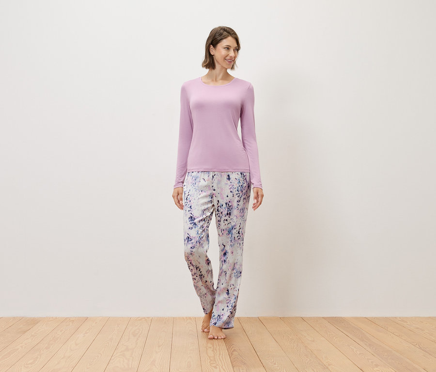 Kobieta stoi boso na drewnianej podłodze, ubrana w fioletowy sweter i wzorzyste spodnie.