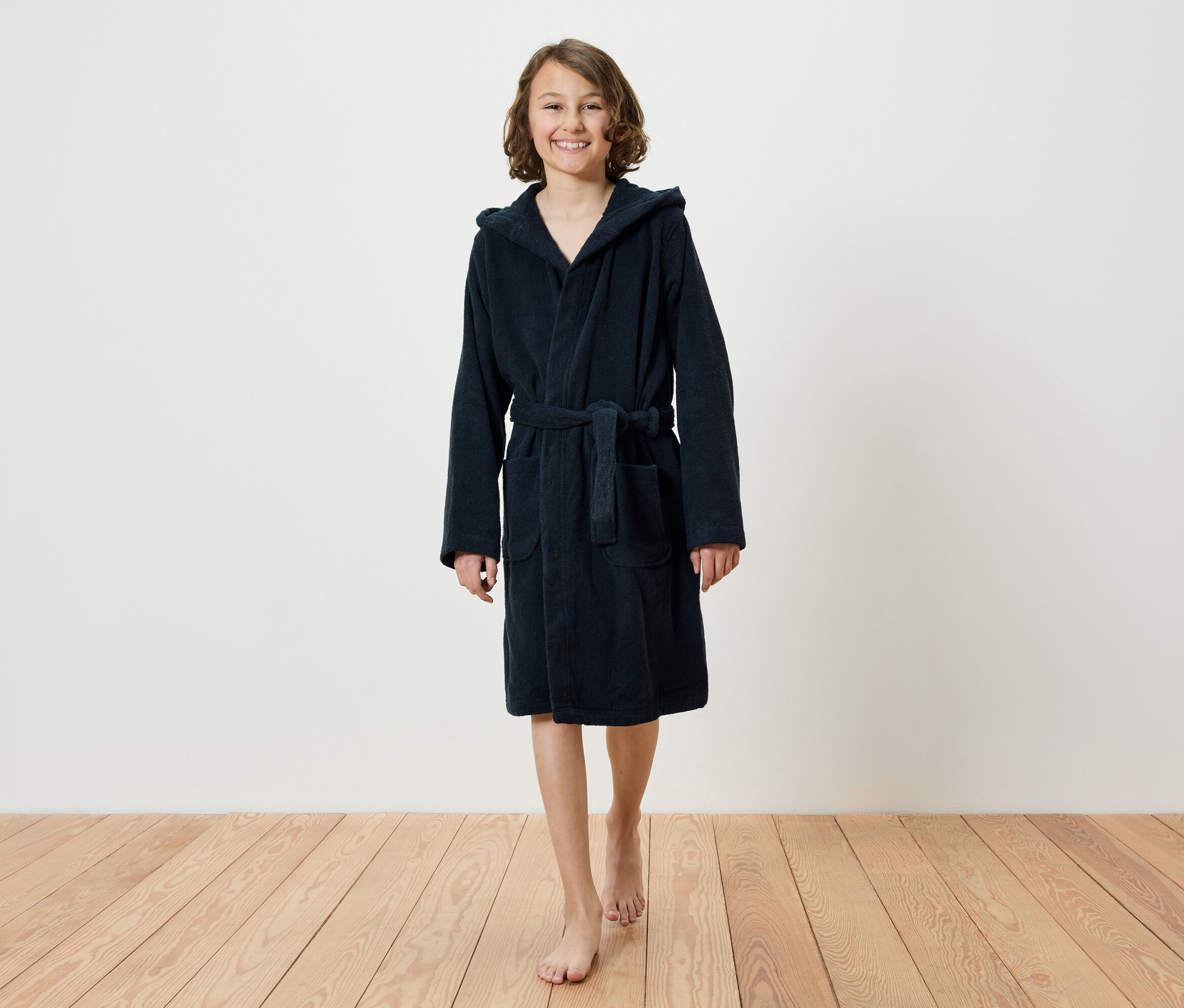 Uśmiechnięta nastolatka idzie boso po drewnianej podłodze, ubrana w granatowy szlafrok z kapturem.