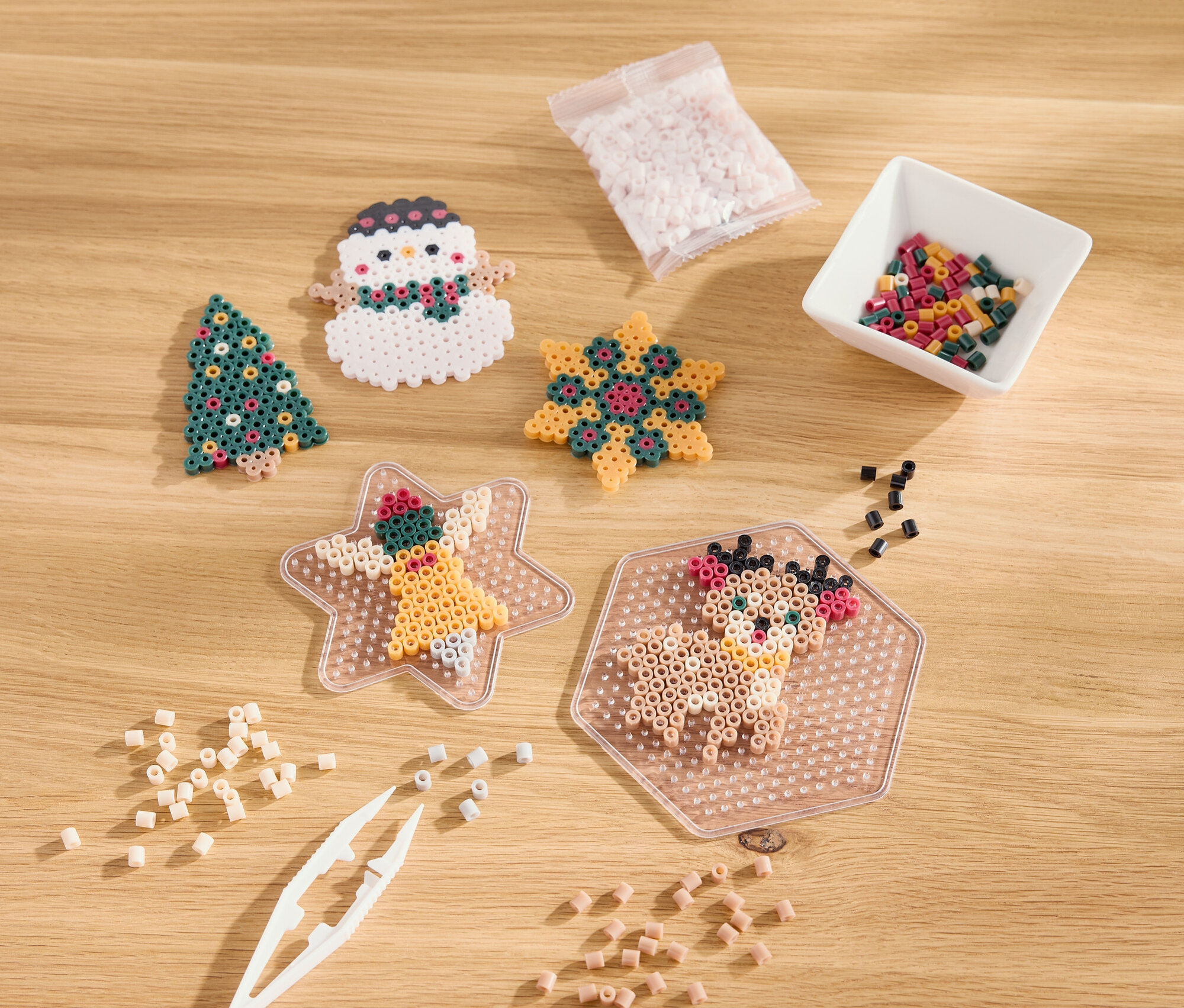 Na drewnianym stole leżą świąteczne ozdoby z koralików, w tym choinka, bałwan, gwiazda i renifer.