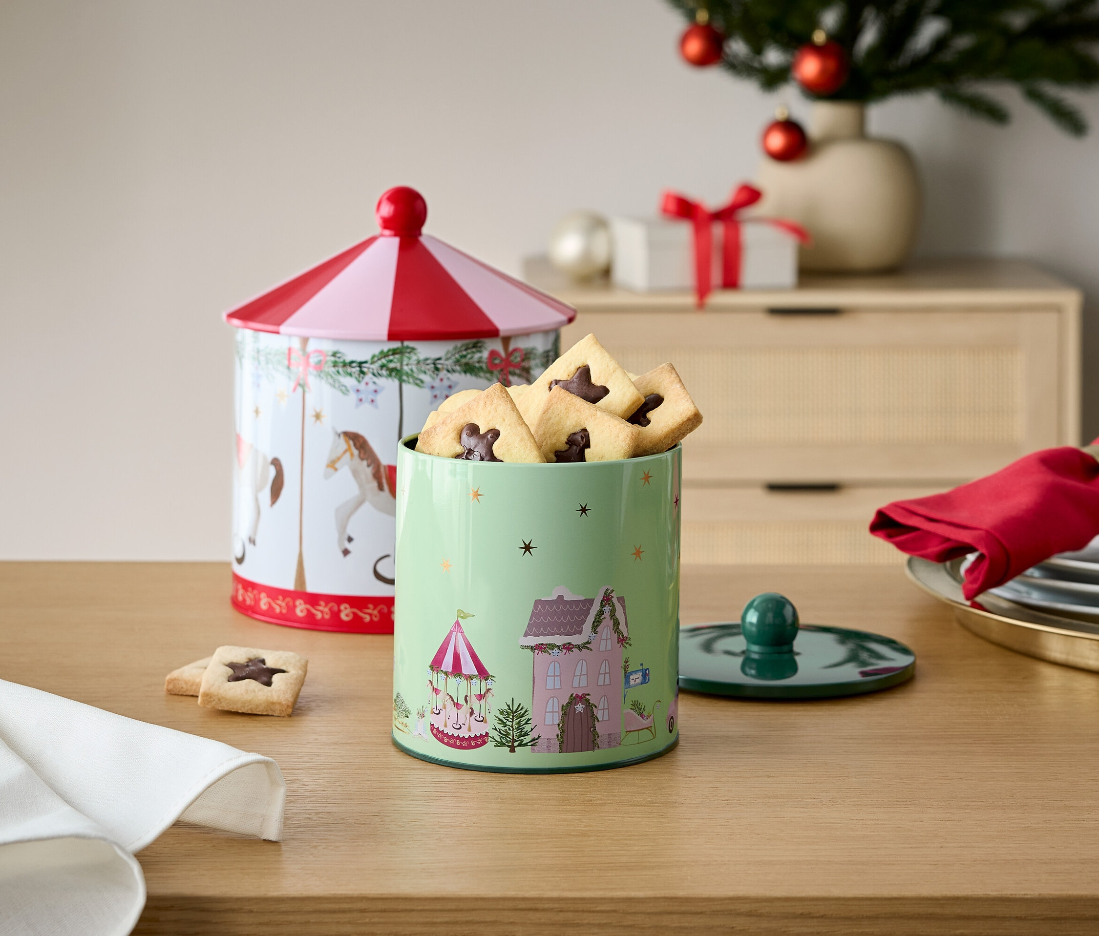 Dwie ozdobne puszki na ciasteczka na drewnianym stole z dekoracjami świątecznymi w tle.