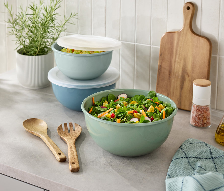 Na blacie kuchennym stoi miska sałaty, drewniane sztućce, solniczka, butelka oleju, deska do krojenia i roślina doniczkowa.