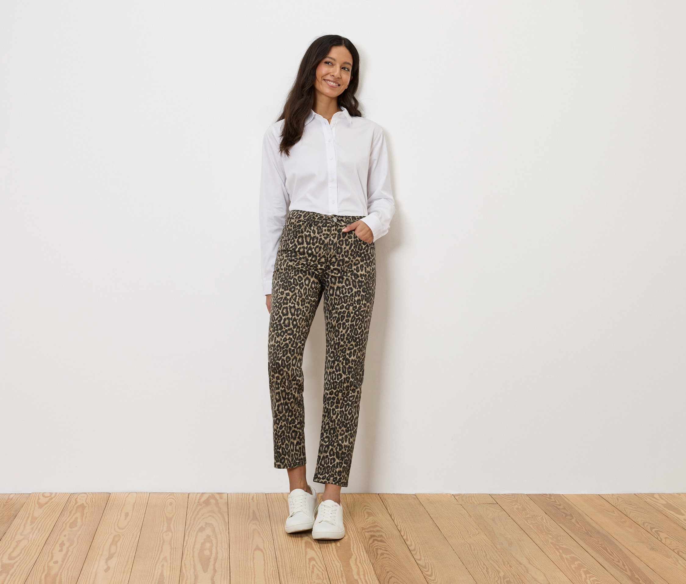 Kobieta w białej koszuli i spodniach w lamparcie cętki, stojąca na tle białej ściany.