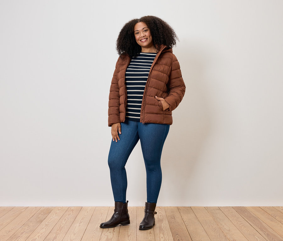 Kobieta z kręconymi włosami pozuje w brązowej kurtce puchowej, niebieskich dżinsach i brązowych butach.