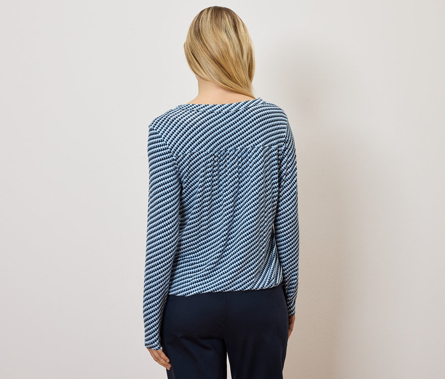 Widok z tyłu kobiety z blond włosami, ubranej w bluzkę w niebiesko-białe paski i granatowe spodnie.