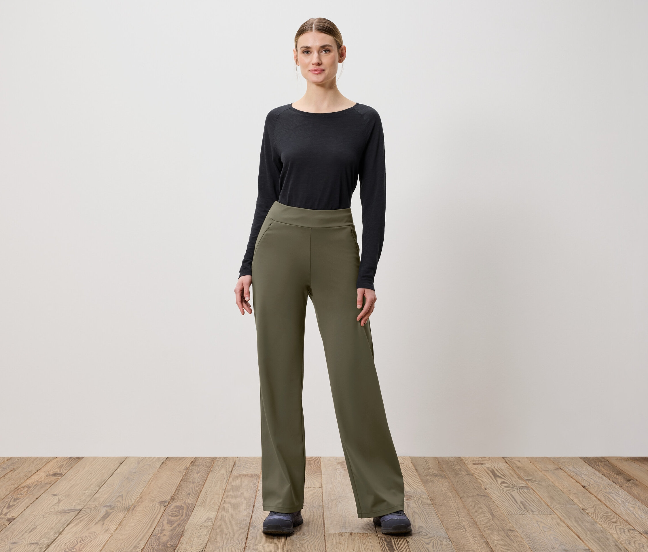 Kobieta pozuje na drewnianej podłodze w oliwkowozielonych spodniach i czarnej bluzce z długim rękawem.