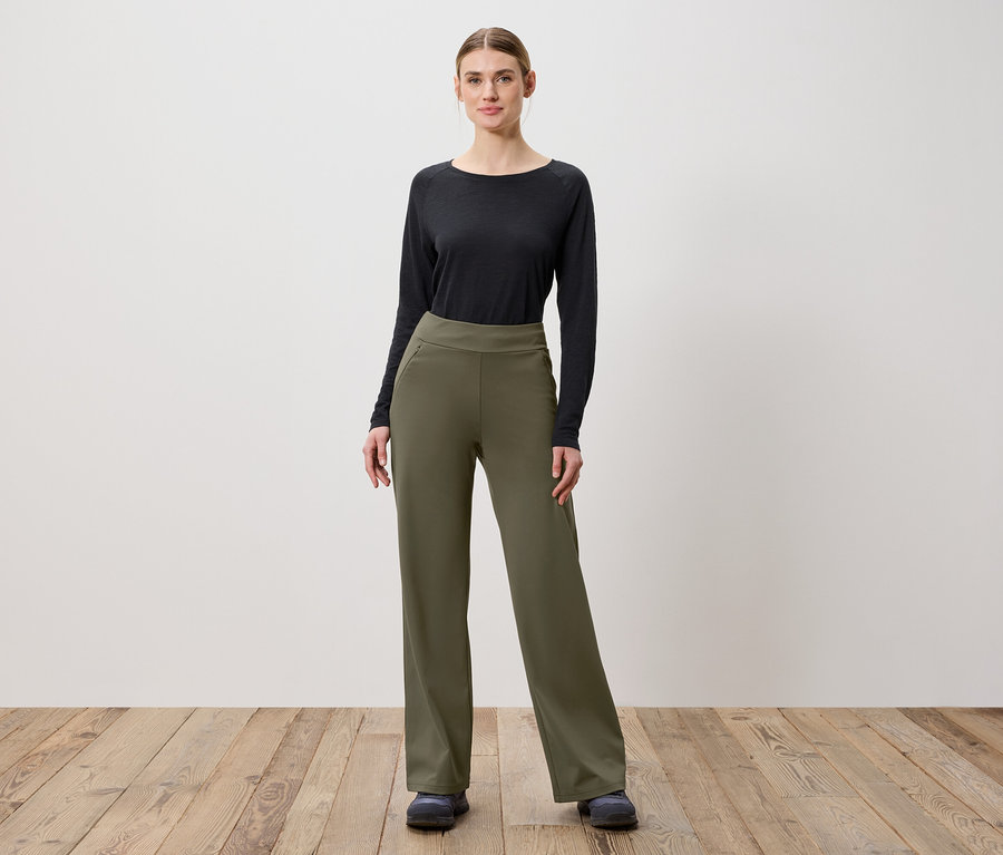 Kobieta pozuje na drewnianej podłodze w oliwkowozielonych spodniach i czarnej bluzce z długim rękawem.