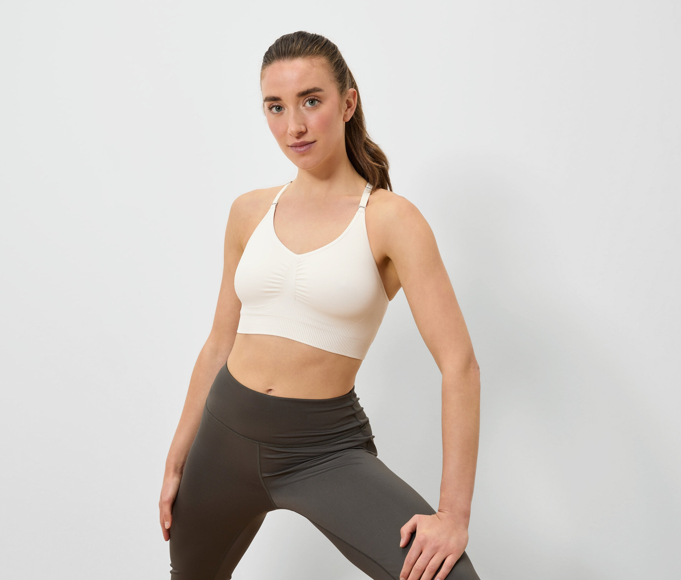 Kobieta pozuje w kremowym staniku sportowym i szarych legginsach na białym tle.