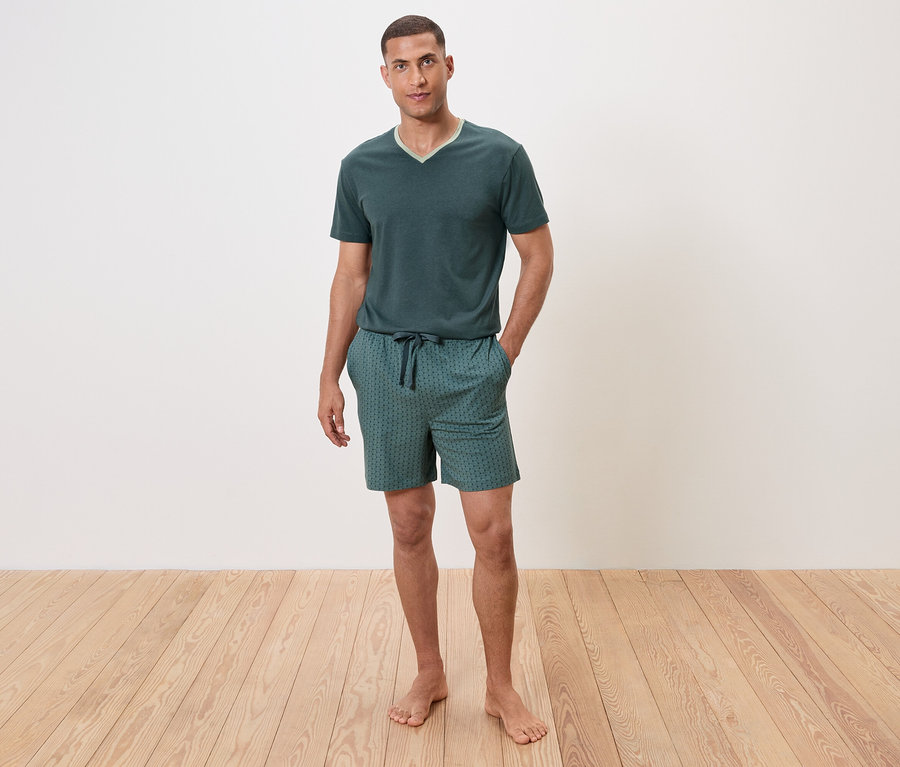 Mężczyzna stoi boso na drewnianej podłodze, ubrany w zieloną piżamę z krótkim rękawem i szorty.