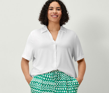 Kobieta stoi z rękami w kieszeniach wzorzystych spodni dżersejowych o długości 3/4 i białej bluzki zapinanej na guziki.