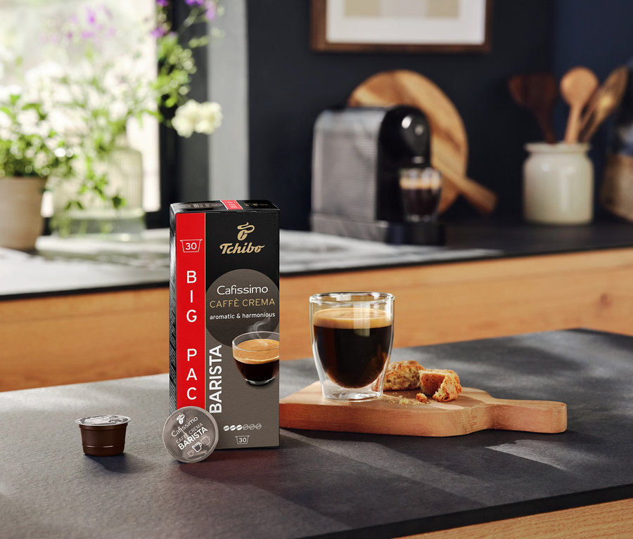 Na blacie kuchennym stoi opakowanie Tchibo Cafissimo Caffè Crema, kapsułka, filiżanka kawy i ciastka.