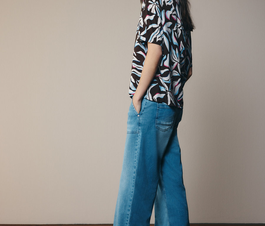 Kobieta w wzorzystej bluzce i dżinsach z szerokimi nogawkami, patrząca w bok.