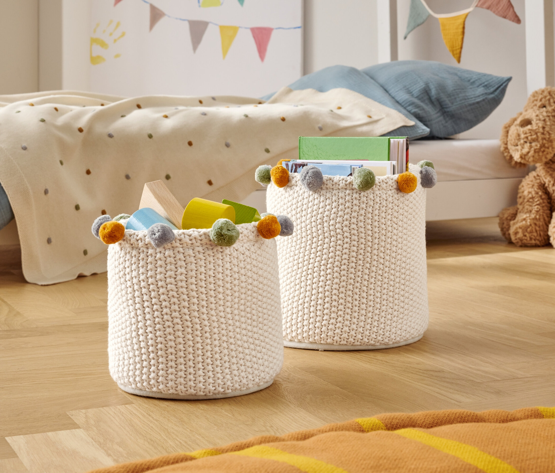 Dwa dziergane kosze do przechowywania z kolorowymi pomponami stoją na drewnianej podłodze w pokoju dziecięcym.