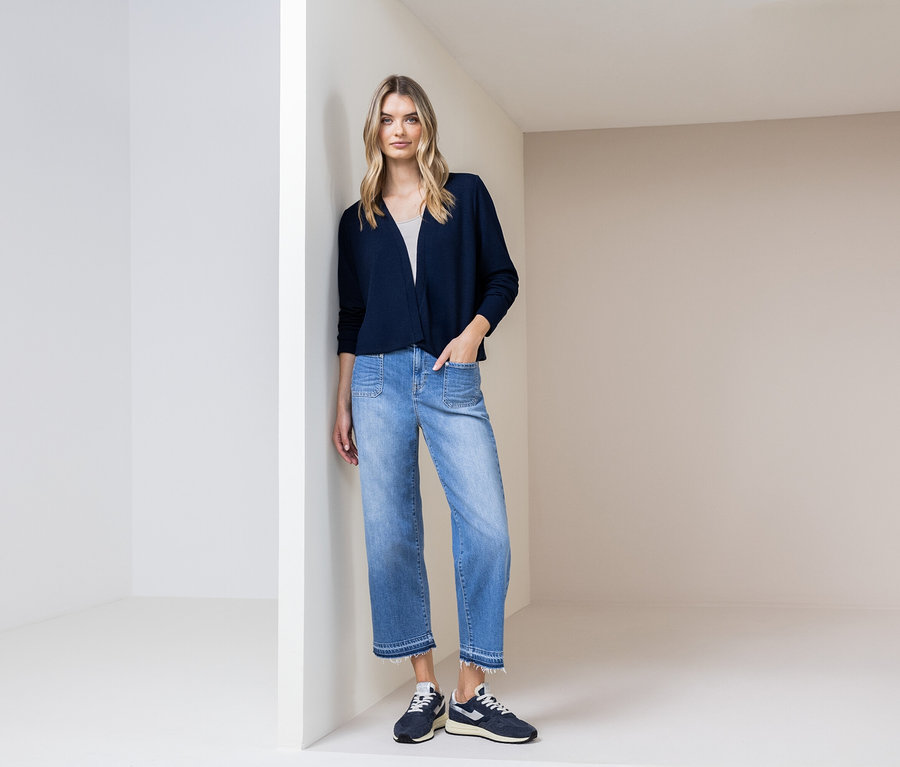Kobieta opiera się o ścianę, ubrana w granatowy kardigan, niebieskie dżinsy i granatowe buty.
