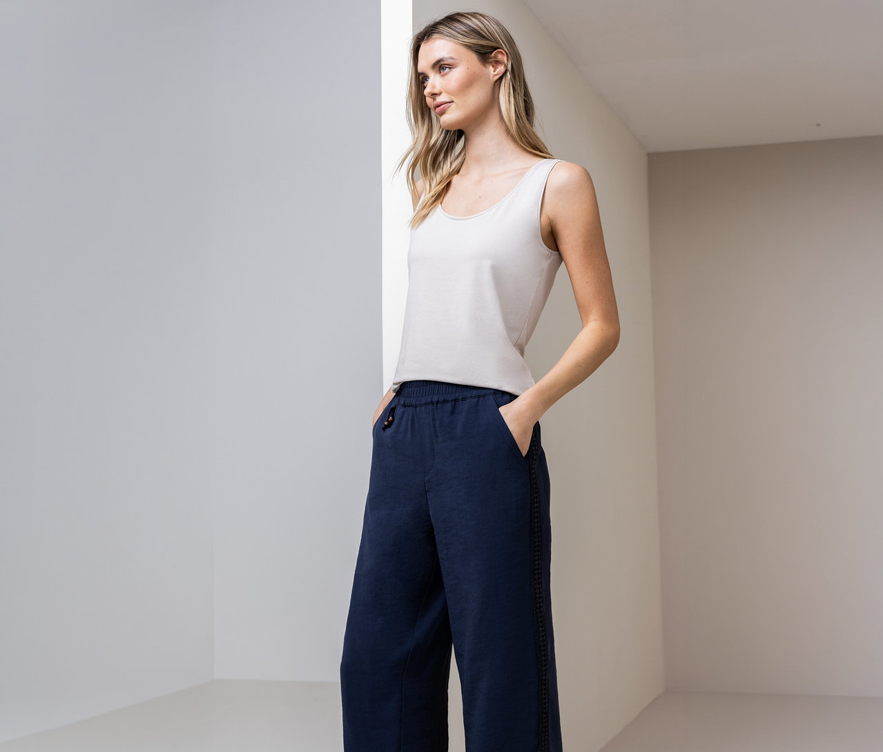 Kobieta w topie basic Street One i niebieskich spodniach z rękami w kieszeniach na tle ściany.