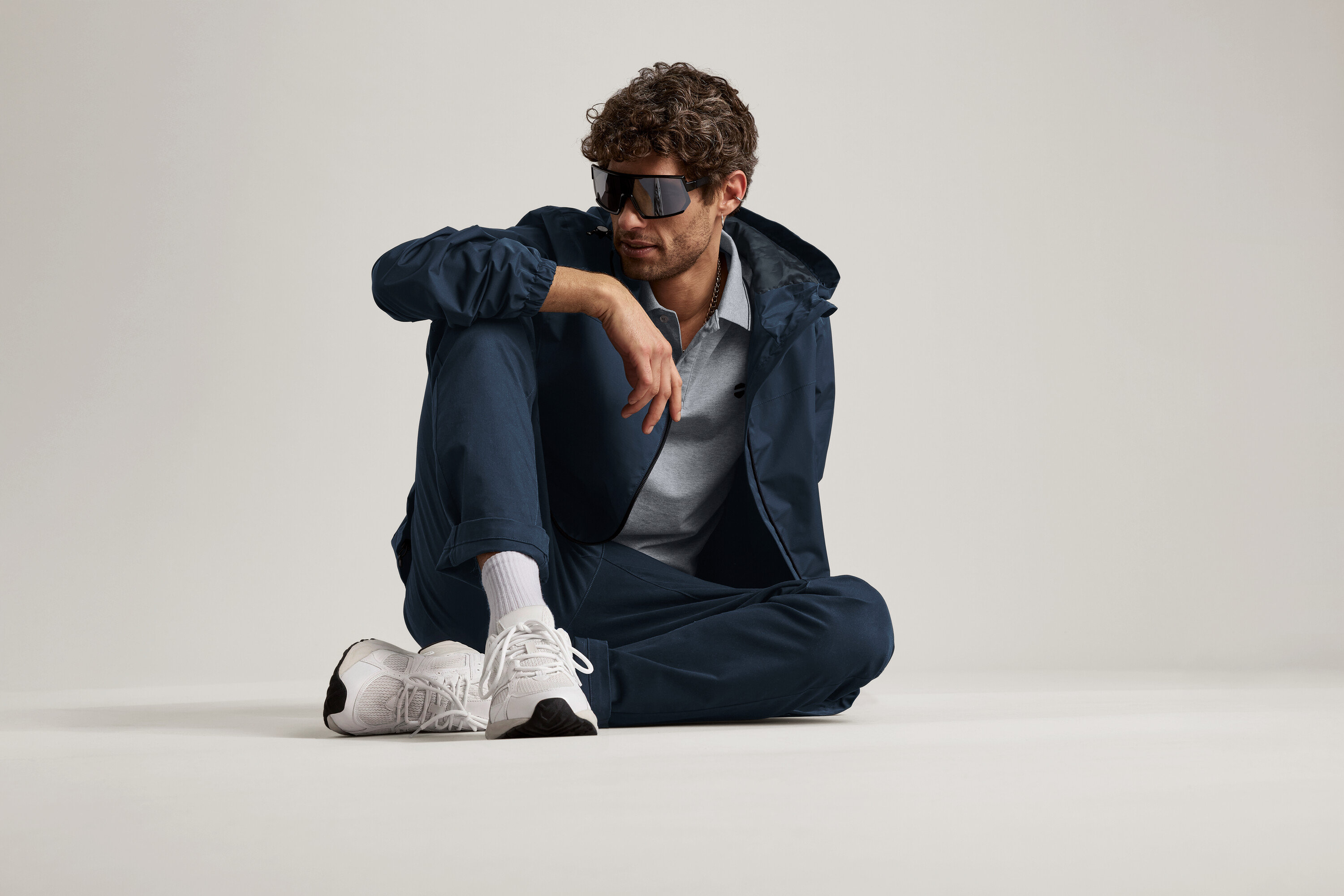 Mężczyzna siedzi na podłodze w granatowych spodniach Chino, kurtce i okularach przeciwsłonecznych.