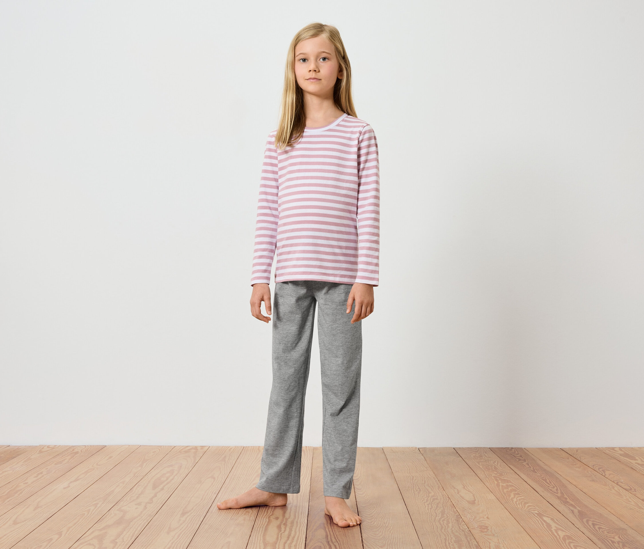 Młoda dziewczyna stoi boso na drewnianej podłodze na tle białej ściany. Ma na sobie bluzkę z długim rękawem w różowo-białe paski i szare spodnie.